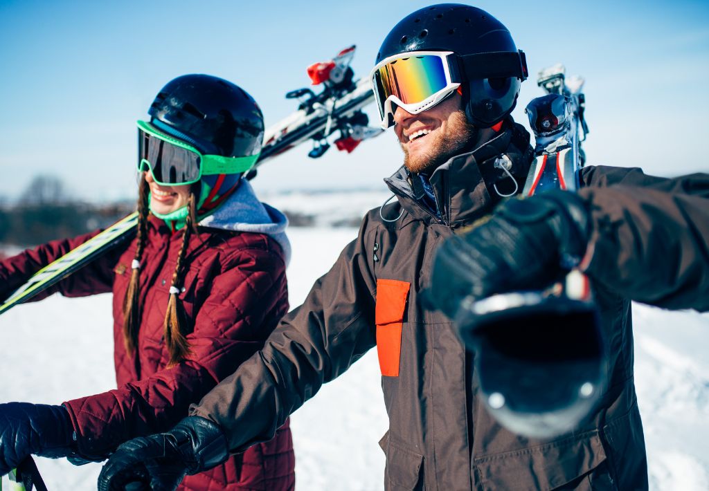 a happy couple is holding their skiis over their shoulders. they are dressed in winter gear and have bright ski goggles and helmets on. they are living their best life in pennsylvania thanks to murrysville functional medicine.