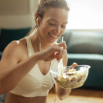 a fit lady is happy, eating a healthy meal in a glass bowl. She is wearing a white sports bra and has earbuds in. This type of semaglutide diet helps people lose the fat while maintaining muscle.