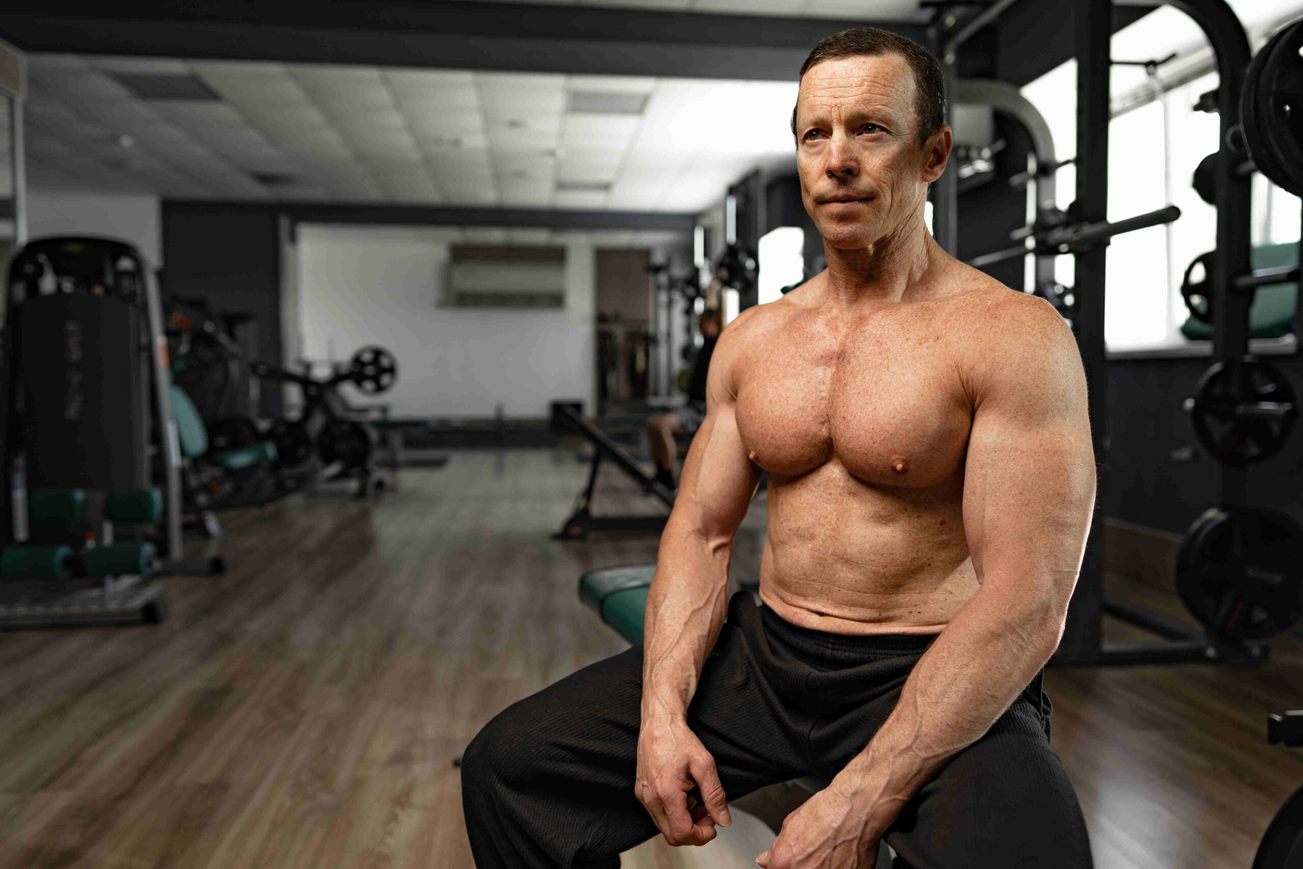 an older guy sits on a bench in the gym without a shirt. He is well built and looks healthier than men half his age. Peptides for men create this kind of effect and he looks great thanks to close medical supervision.